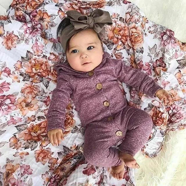 Baby wearing a purple hooded romper with brown buttons, lying on a floral blanket.