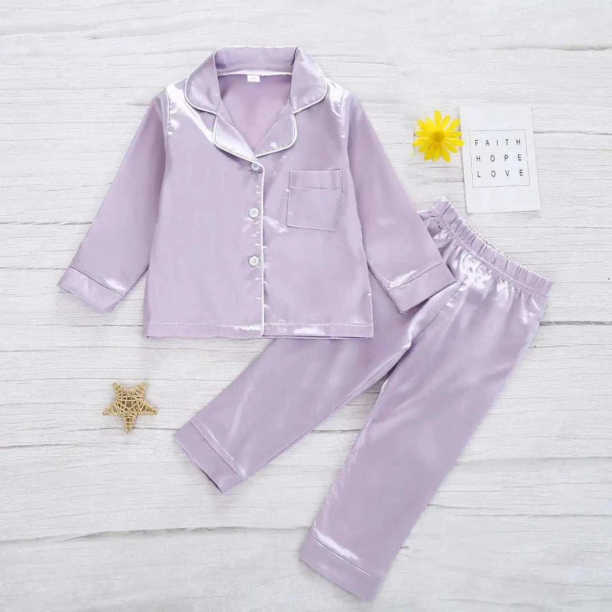 Luxurious silk satin toddler pajamas in light purple, long-sleeved top with buttons and matching pants, laid on a light background.